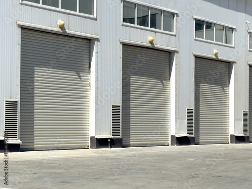 Three closed grey roller doors line the exterior facade of a modern industrial warehouse building. Commercial logistics, storage facility, business infrastructure.
