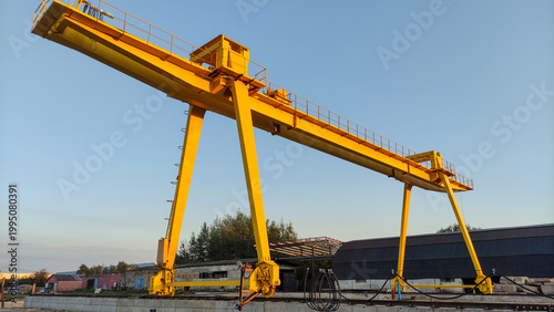 A massive yellow gantry crane stands over an industrial railway yard under a clear blue sky. Heavy logistics operation, freight transportation, industrial infrastructure.