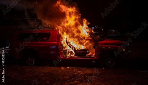 A red vehicle is on fire in a dark area. Flames engulf the inside and outside of the vehicle. Smoke billows into the night sky as the fire burns brightly in the darkness.