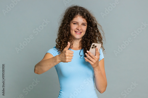 Woman scrolling news feed on smartphone smiling warmly giving thumbs up like gesture with pleased positive reaction. Girl on gray background shows appreciation of favorite online content happily