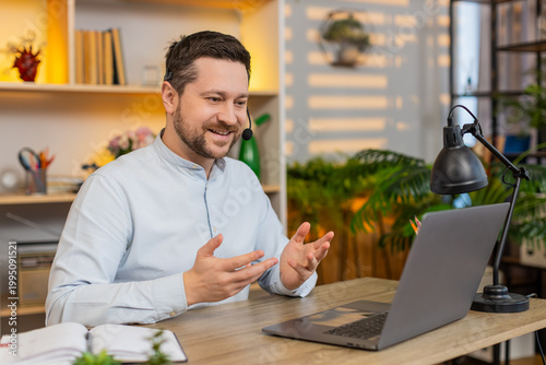Young man on home table smiles in headset and listens to client on laptop while answering polite support call. Adult guy on home table works as remote operator giving help and service online each day.