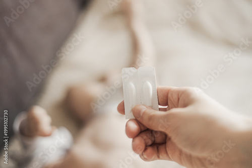 Close-up of a hand holding a package of two suppositories against the background of a lying baby at home