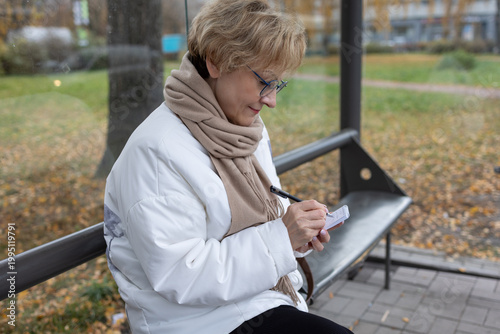 Thoughtful female in white jacket and beige scarf writing notes while standing by metal bench in fall setting.