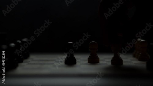 Cinematic close-up of a hand moving a chess piece in a dark atmospheric room. Low-key lighting highlights strategy, planning, and intellectual competition. Dramatic silhouette of a chess game.