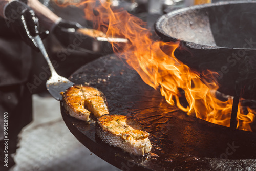 Salmon steaks grilling on large metal fire pit surface. Chef preparing fish steak over open flame. Gourmet street food cooking process in outdoor kitchen setting.
