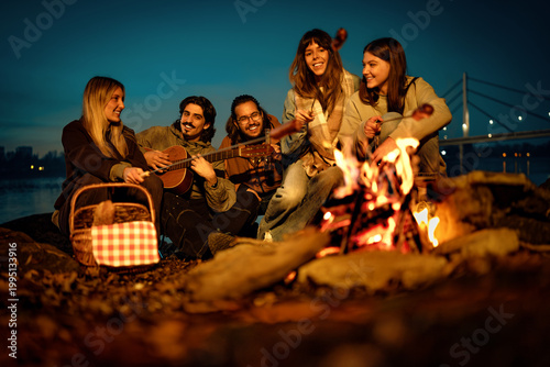 Friends gather around a campfire by a river while playing music and enjoying a picnic during the evening