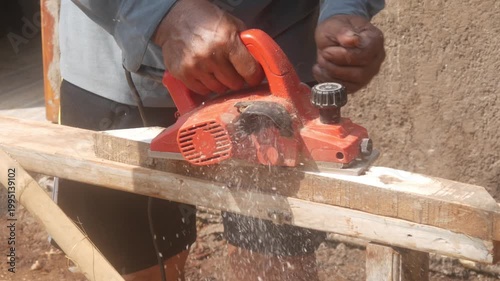 A woodworker uses an electric planer to smooth the surface of a wooden beam on a building construction project.