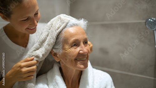 A smiling caregiver providing hygiene assistance by drying a happy senior elderly woman's hair with a towel in a bathroom, focusing on compassionate home care.