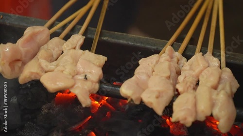 Chicken satay skewers grilling over smoky charcoal, raw marinated meat cooking on open fire in an Asian street food BBQ scene.
