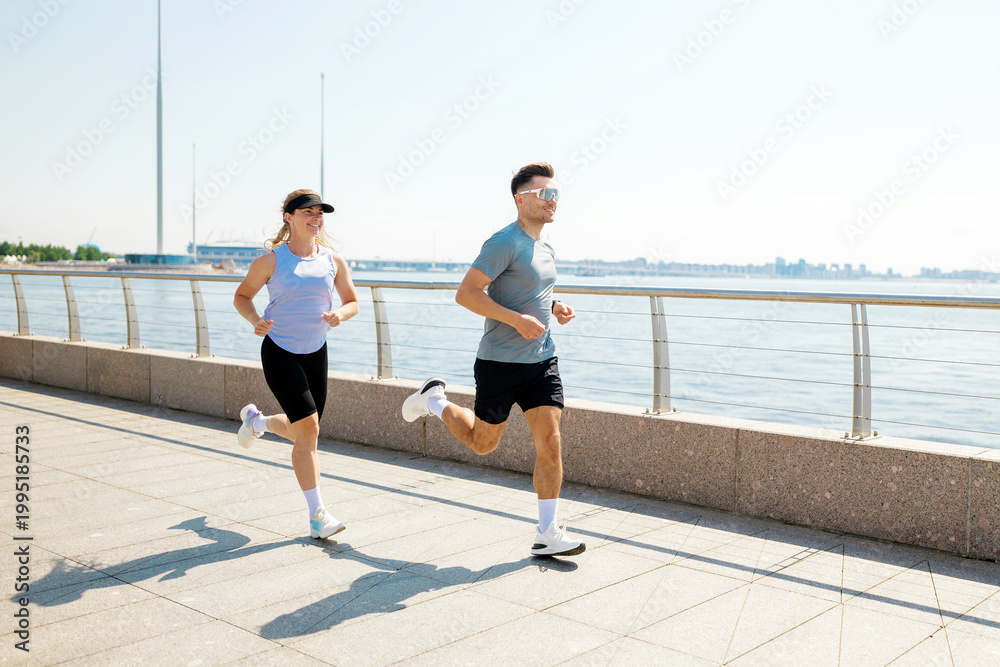 Fototapeta premium Joyful couple runs along the waterfront path, embracing sunlight on a bright day in a vibrant outdoor setting