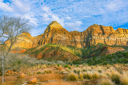 Tablou pe pânză Golden sunlight illuminates the layered red and orange sandstone cliffs of Zion National Park above a dry scrubland valley under a partly cloudy sky