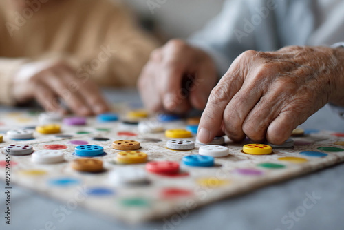 Two hands gently place colorful buttons on a game board. Older skin shows wrinkles and care. The game suggests quiet, thoughtful play. Warm tones evoke comfort and connection
