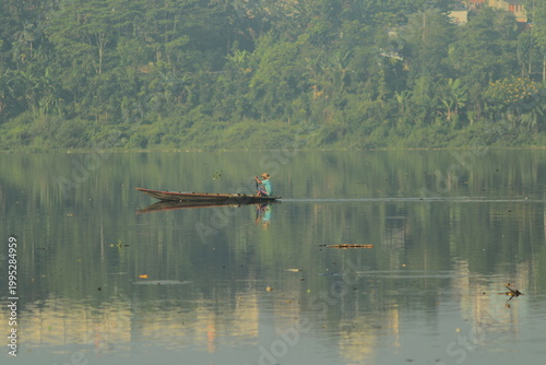 Man rowing traditional canoe on tranquil lake with mirror reflections, tropical countryside scenery with greenery, calm water surface and quiet morning mood