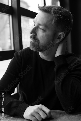 Black and white portrait of a man in his 40s seated by a restaurant window in Ottawa, Canada. Thoughtful side profile with soft natural light, casual sweater, and calm modern atmosphere indoors.