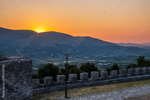 A warm sunset from Berat Castle walls frames the city, Osum River valley, and distant mountains with an orange glow. Historic stone wall visible in Berat, Albania.