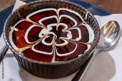 A rich, dark brown trilece dessert with caramel, presented in a ribbed cup with a decorative white cream swirl pattern, resembling a flower in Tirana, Albania.