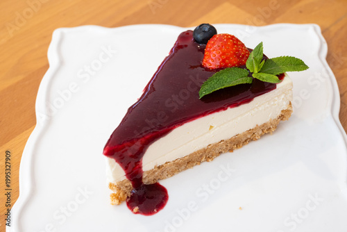 A decadent dessert slice of creamy cheesecake featuring a vibrant red berry topping, fresh strawberry, blueberry, and mint garnish on a white plate.