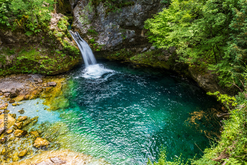 An elevated wide shot captures the vibrant emerald-blue pool of the Blue Eye of Theth, fed by a waterfall, with a varied stream bed and rocky banks in Theth, Albania.