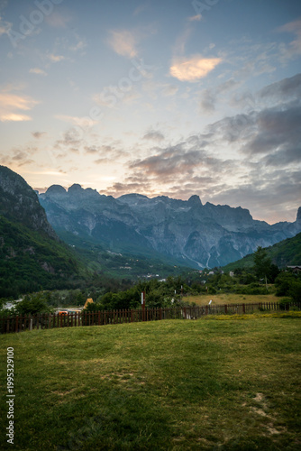 A vertical panorama captures a verdant green field and wooden fence foregrounding a dramatic mountain valley under a cloudy sunset sky, in Theth, Albania.