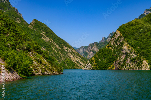 Expansive view of the Koman Lake gorge, showcasing lush green mountains rising dramatically from tranquil turquoise water under clear daylight. Pristine natural environment.