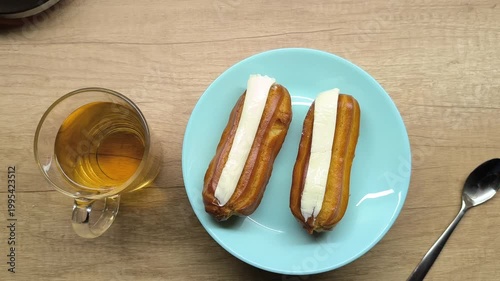 Two eclairs lie on a turquoise plate on a wooden table next to a cup of tea and a napkin, close-up, exquisite dessert, homemade cream cakes, tea party.
