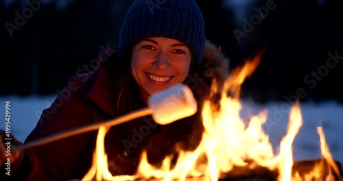 A smiling young woman roasting marshmallows over a cozy outdoor campfire during a winter evening.