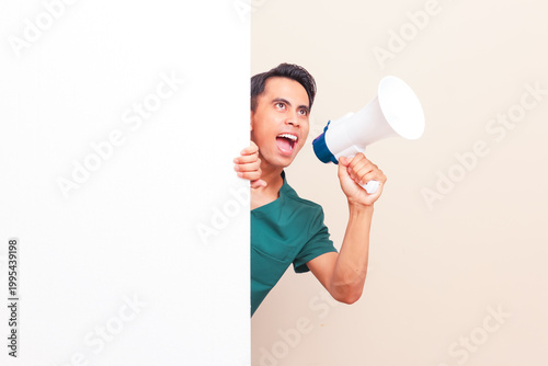 Canvastavla Excited Southeast Asian male nurse or doctor in green scrubs peeking from behind a large blank white billboard while holding a megaphone making an announcement with copy space on isolated background