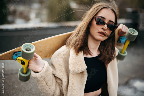 Young brunette in sunglasses posing on roof with skateboard