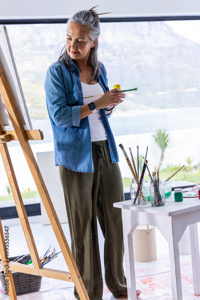 Naklejka premium Senior woman standing at wood easel, holding brush and jar, painting on canvas in coastal studio