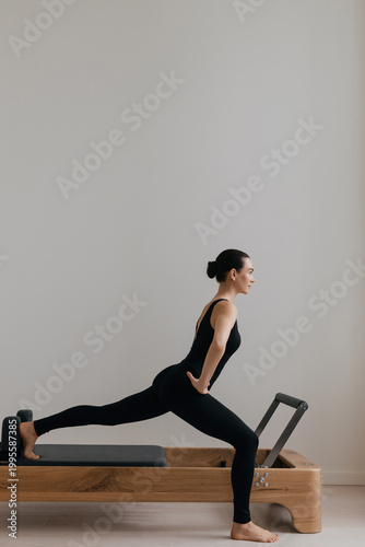 Pilates reformer machine used by fit woman in black leotard against white wal