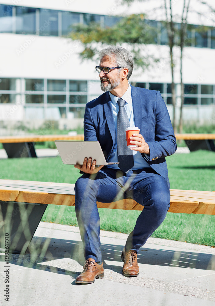Obraz premium portrait of a senior busninessman using a laptop in park in front of corporate buildings
