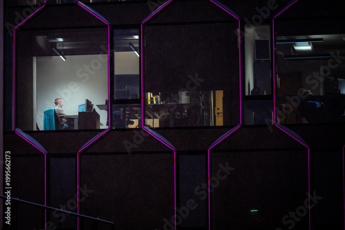 Tallinn, Estonia - 03 January 2025: View of a solitary figure bathed in warm light within a stark, dark modern building, its geometric windows outlined in vibrant magenta against the night.