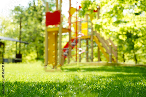 A wide-angle shot of a scuffed soccer ball sitting on a lush green lawn, with a colorful children's playground and a dense green forest blurred in the background under bright, natural light.