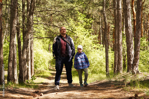 A father and his young son stand together on a dirt path in a sun-drenched pine forest, looking ahead during a family hike or nature walk.