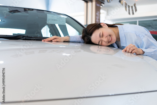 Happy Asian woman hugging her new car in a showroom. Successful vehicle purchase, car ownership, and extreme customer satisfaction concept.