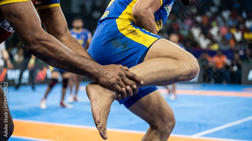 Kabaddi match action showing ankle hold tackle, professional athletes competing in indoor stadium, traditional Indian contact sport tournament
