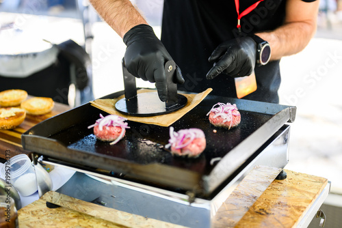 Chef preparing smash burgers on a flat top grill, adding toppings to freshly cooked beef patties with melted cheese, capturing street food cooking process and professional burger preparation in a