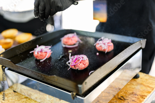 Chef preparing smash burgers on a flat top grill, adding toppings to freshly cooked beef patties with melted cheese, capturing street food cooking process and professional burger preparation in a