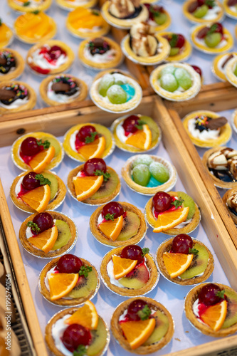 Assorted dessert cups with fruit and chocolate displayed in market stall, colorful sweet selection with orange kiwi grapes and mango, ready for sale in street food setting