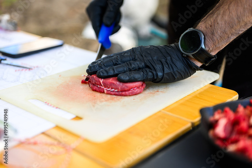 Close-up of a chef in black nitrile gloves precisely trimming a thick-cut ribeye steak with a sharp knife on a cutting board during a professional Steak Cookoff Association (SCA) BBQ competition.