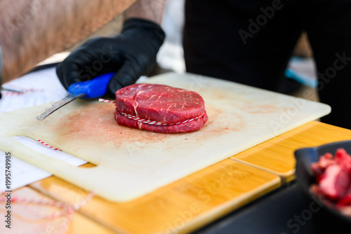 Close-up of a chef in black nitrile gloves precisely trimming a thick-cut ribeye steak with a sharp knife on a cutting board during a professional Steak Cookoff Association (SCA) BBQ competition.