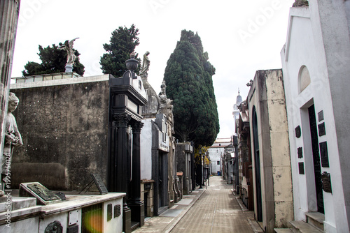A street-level perspective of a narrow lane in Recoleta Cemetery, Buenos Aires. The path is lined with diverse, towering mausoleums made of granite and marble, featuring elaborate sculptures, crosses
