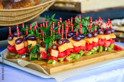 Rows of colorful fresh fruit skewers featuring grapes, melon, watermelon, and kiwi, garnished with fresh mint and presented on a wooden board at a catered event or healthy breakfast buffet.