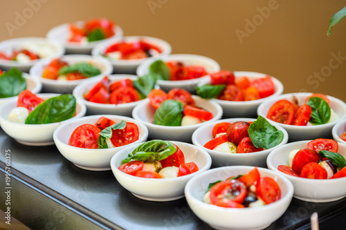 Rows of individual ceramic bowls filled with mini Caprese salad appetizers, featuring fresh cherry tomatoes, mozzarella pearls, and basil leaves drizzled with balsamic glaze at a catered event.
