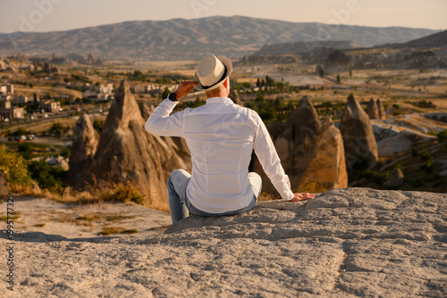 Photos Young man in straw hat and white shirt sitting on a rock overlooking Cappadocia valley in Turkey