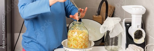 A woman stands in her kitchen, mixing cabbage for fermentation. She uses a spoon to stir the cabbage while readying jars for storage. The atmosphere is filled with food preparation, banner