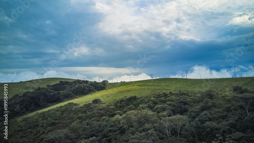 Aerial view of rolling emerald hills crowned with a dense, dark green canopy under a dramatic expanse of white and deep blue clouds, Obudu, Cross River, Nigeria.