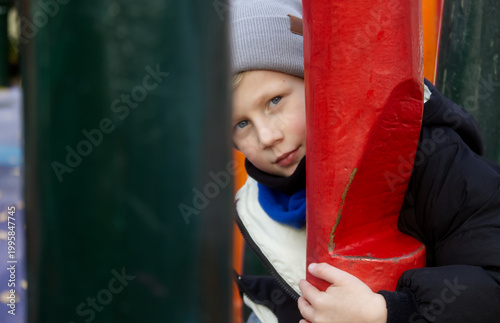 Cute blond boy playing hide and seek behind a red pole on a playground
