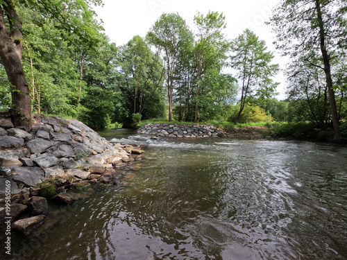 Bild auf Leinwand Flowing river stream with stone riprap bank stabilization in green summer forest landscape