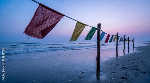 A line of colorful, tattered prayer flags strung across wooden posts on a quiet beach. The scene is captured at twilight, with soft purple and blue hues reflecting on the wet sand and ocean waves.
